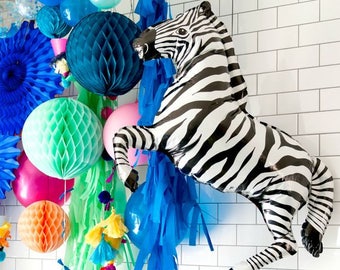 May include: A zebra balloon in black and white stripes hangs from a ceiling with blue and green tissue paper streamers. A table is set with a cake, donuts, and a pitcher of lemonade. The table is decorated with blue and white striped paper cups and confetti.