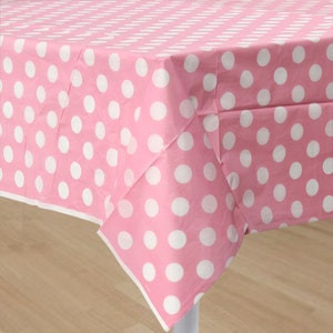 Puede incluir: Un mantel rosa con un patrón repetitivo de grandes lunares blancos. El mantel está colocado sobre una mesa, con los bordes visibles. La tela parece ser un material ligero, adecuado para fiestas o cenas informales.
