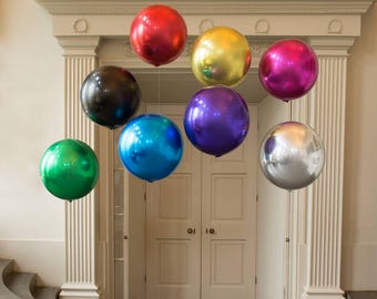 Puede incluir: Nueve globos grandes, redondos y metálicos de varios colores, incluyendo rojo, dorado, magenta, morado, azul, verde y plateado, están suspendidos del techo en una habitación con paredes blancas y una puerta blanca. Los globos están dispuestos en forma de semicírculo.
