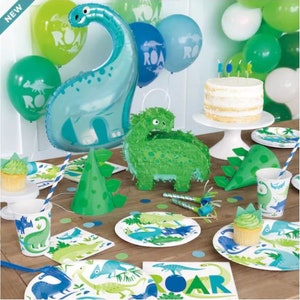 May include: A dinosaur-themed party table setting with green and blue decorations. There is a blue dinosaur balloon, green and blue paper plates, green and blue napkins, a green dinosaur pi&ntilde;ata, and a white cake with candles. The table is covered in a brown tablecloth.