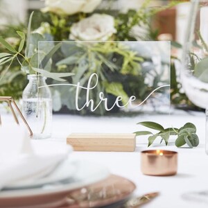 Op de afbeelding: Een helder acryl tafelnummer met het woord "three" in elegant wit script. Het nummer wordt weergegeven op een klein houten blok, met een koperkleurige kaars en groen in de achtergrond.