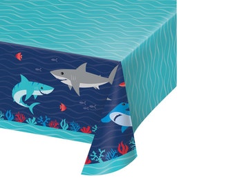Puede incluir: Mantel de mesa de plástico azul y blanco con temática de tiburones. El mantel presenta un fondo azul océano con olas blancas y un borde de coral rojo y azul. Hay tres tiburones en el mantel, dos tiburones azules y un tiburón gris.
