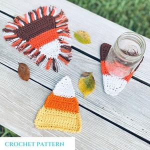 May include: Three crocheted coasters in the shape of candy corn. The coasters are orange, yellow, and white. The coasters are on a wooden table with fall leaves.