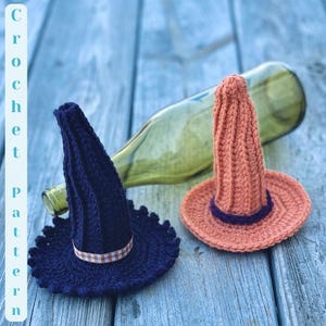 May include: Two crocheted witch hats, one in dark blue with a white and red gingham ribbon and the other in orange with a purple ribbon. The hats are sitting on a wooden surface.