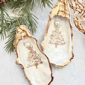 May include: Two white oyster shell ornaments with gold trim, each featuring a gold-painted Christmas tree and a pearl accent. The ornaments are hanging from a gold wire.