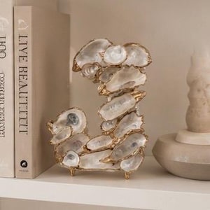 May include: A decorative letter "J" made of oyster shells with gold-colored accents. The sculpture sits on a white shelf next to a book with the text "LIVE BEAUTIFUL". The shells are white and iridescent.