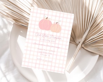 May include: A pink and white gingham patterned invitation card with two pumpkin illustrations. The text reads "Our Little Pumpkin is turning one!" with event details. The card is on a white plate with a dried palm leaf.