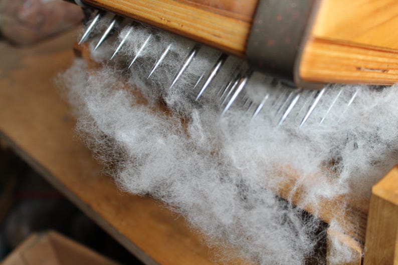Puede incluir: Primer plano de un cardador de lana de madera con un peine de metal y fibras de lana blancas en proceso.