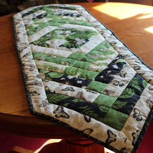May include: A green and white quilted table runner with a geometric pattern and butterfly print. The runner has a dark blue trim.
