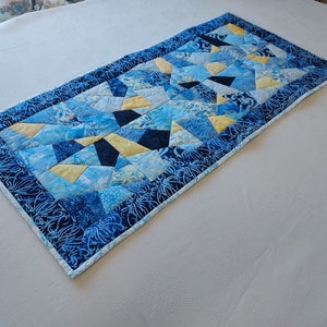 May include: A blue and yellow patchwork table runner with a geometric design. The runner is made of fabric with a floral pattern and has a white border.