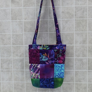 May include: A purple and green patchwork tote bag with a long strap. The bag is made of different fabrics with floral and geometric patterns.