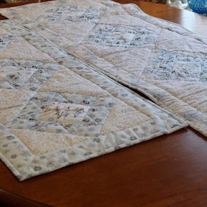 May include: Two white and blue quilted table runners with a diamond pattern. The runners are made of fabric scraps in various shades of blue and white.