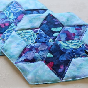 May include: Two blue and purple quilted fabric placemats with a floral pattern. The placemats are hexagonal in shape and have a light blue background with a darker blue floral pattern.