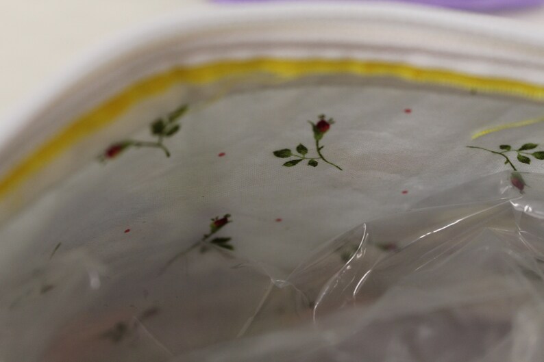 May include: A white fabric bag with a yellow zipper and a floral pattern. The fabric has a sheer, translucent quality and features small red flowers and green leaves.