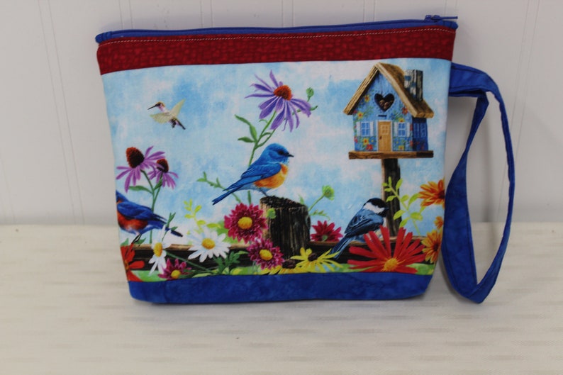 May include: Blue zippered pouch with a blue bird, a birdhouse, and flowers on a blue and white background. The pouch has a red and blue trim and a blue handle.