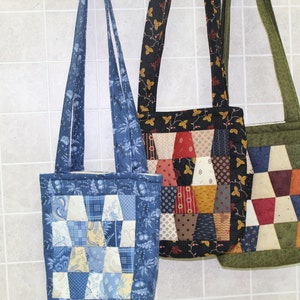May include: Three quilted tote bags hanging on a white tiled wall. The bag on the left is blue with a white and yellow patchwork design. The middle bag is black with a brown, red, and white patchwork design. The bag on the right is green with a brown, red, and white patchwork design.