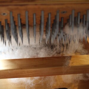 Puede incluir: Un marco de madera con una fila de agujas met&aacute;licas, utilizado para cardar la lana. Las fibras blancas est&aacute;n siendo procesadas por las agujas.