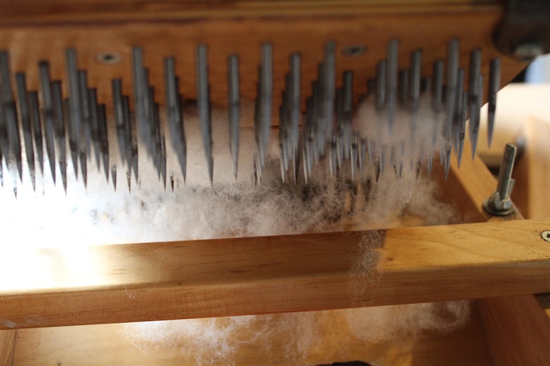 Puede incluir: Un marco de madera con una fila de agujas met&aacute;licas, utilizado para cardar la lana. Las fibras blancas est&aacute;n siendo procesadas por las agujas.