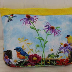 May include: A yellow zippered pouch with a blue sky and floral print. The print features purple flowers, yellow and blue birds, and green leaves.