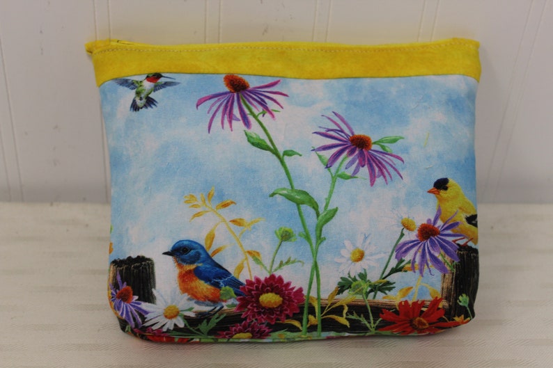 May include: A yellow zippered pouch with a blue sky and floral print. The print features purple flowers, yellow and blue birds, and green leaves.