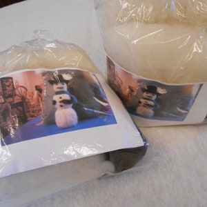 May include: Two clear plastic bags containing white fiberfill and a brown fiberfill. Each bag has a printed image of a snowman with a black hat and a blue scarf. The image also includes a wooden spinning wheel and other craft supplies.