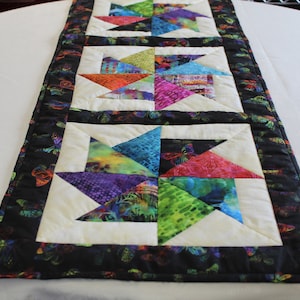 May include: A colorful table runner with a black background and a white border. The runner features a repeating pattern of colorful pinwheels with a butterfly design on the border.