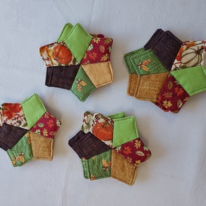 Puede incluir: Conjunto de seis posavasos acolchados en forma de estrella con temática otoñal. Los posavasos presentan una variedad de telas en tonos marrones, verdes, rojos y dorados con patrones de calabazas, hojas y bellotas.