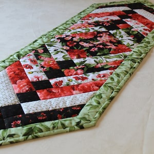 May include: A green, black, white, and red quilted table runner with a floral pattern. The runner has a diamond shape and is made of fabric.