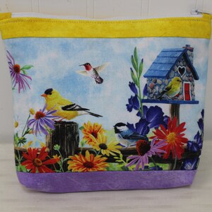 May include: A purple and yellow zippered pouch with a birdhouse and birds on a floral print. The birdhouse is blue with a white heart on the front. The birds are yellow, blue, and red.