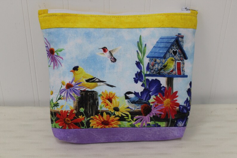 May include: A purple and yellow zippered pouch with a birdhouse and birds on a floral print. The birdhouse is blue with a white heart on the front. The birds are yellow, blue, and red.