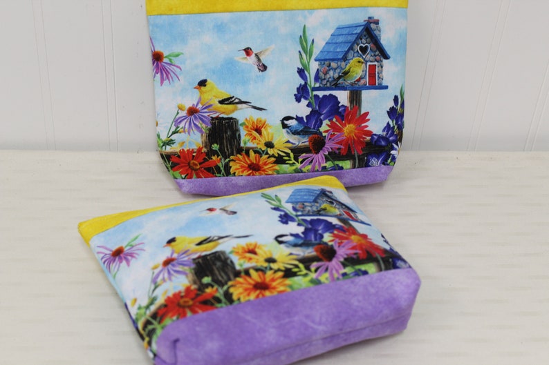 May include: Two small zippered pouches with a floral and bird print. The pouches are lined with purple fabric and have a yellow trim. The print features a birdhouse, birds, and flowers.