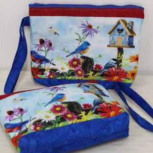 May include: Two blue zippered pouches with a floral and bird design. The pouches have blue handles and a red and blue trim. The design features bluebirds, a birdhouse, and flowers.