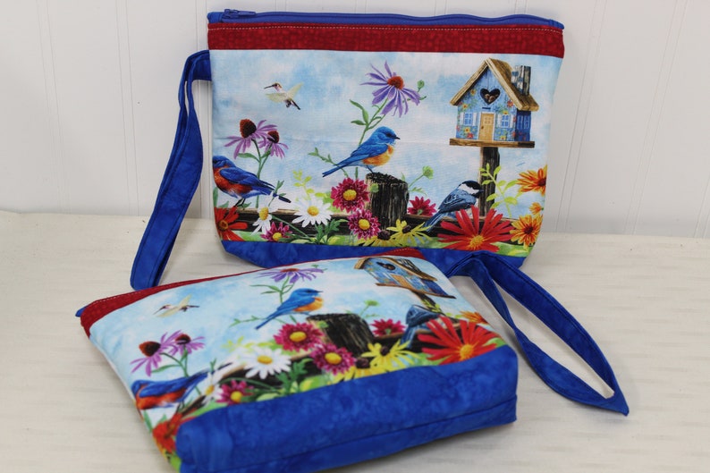 May include: Two blue zippered pouches with a floral and bird design. The pouches have blue handles and a red and blue trim. The design features bluebirds, a birdhouse, and flowers.
