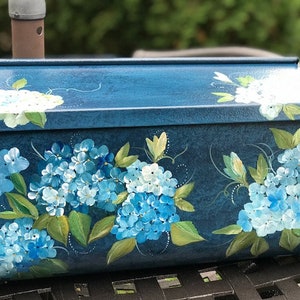 May include: A blue mailbox featuring a hand-painted design of blue and white hydrangeas. The mailbox has a textured surface and a slightly curved top.
