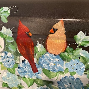May include: A hand-painted mailbox with a black background and a white picket fence. Two red cardinals are perched on the fence, surrounded by blue and green flowers.
