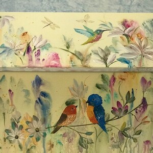 May include: A yellow wooden box with a hinged lid, painted with a whimsical floral design. The lid features a hummingbird in flight, while the box shows two birds perched on a branch, surrounded by flowers and leaves.
