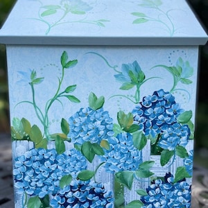 May include: A light blue wooden box with a white picket fence and blue hydrangeas painted on the front. The box has a hinged lid.