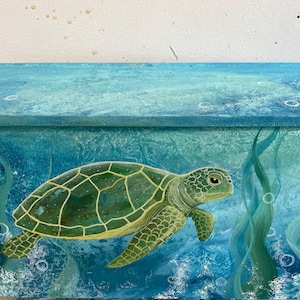 May include: A hand-painted wooden box featuring a green sea turtle swimming in blue water with bubbles and seaweed. The box has a textured, ocean-themed design with shades of blue and green.