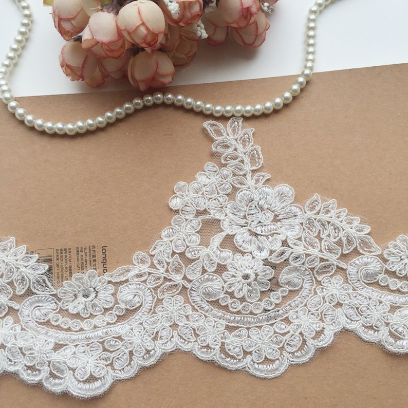 May include: Ivory lace trim with floral and scrollwork embroidery. The delicate trim features a scalloped edge and is embellished with small, sparkling accents. A string of pearls and pink flowers are in the background.
