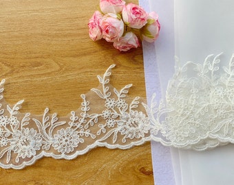 May include: Ivory floral lace trim with a scalloped edge, featuring embroidered flowers and leaves. The lace is displayed on a wooden surface with a bouquet of pink roses and white fabric in the background.
