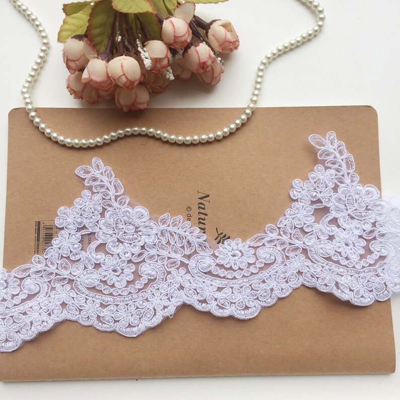 May include: White floral lace trim with a scalloped edge. The lace features detailed floral and leaf patterns. A string of pearls and a bouquet of pink flowers are also in the image.