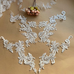 May include: Four white lace appliques with floral and scroll designs. The appliques are arranged in a symmetrical pattern on a brown background.