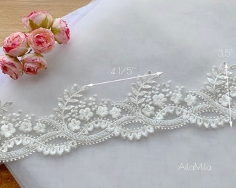 May include: White floral embroidered lace trim, 4 1/5 inches wide and 3.5 inches high, with delicate flower and leaf patterns. The trim is displayed on a white fabric background, with a small bouquet of pink roses in the upper left corner.