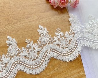 May include: White floral lace trim with a scalloped edge. The lace features detailed embroidered flowers and leaves. A bouquet of pink roses rests on a wooden surface near the lace.