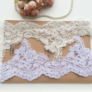 May include: Close-up of white floral lace trim. The lace features a delicate, scalloped edge and intricate floral patterns. The trim is layered on a brown surface, with a string of pearls and flowers in the background.