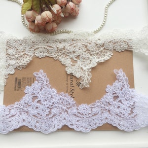 May include: Two white lace trims with floral and leaf patterns are displayed. One is off-white, the other is bright white. The lace features detailed floral designs and scalloped edges. A string of pearls and pink flowers are in the background.
