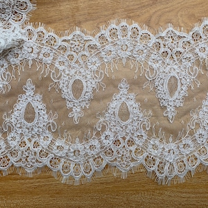 Może przedstawiać: Biała koronka z delikatnym wzorem kwiatowym. Koronka ma falowany brzeg i idealnie nadaje się do dodania elegancji każdemu projektowi.