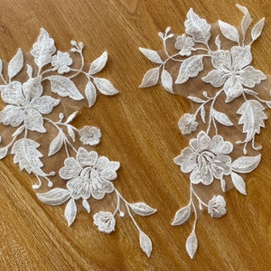 May include: Two white floral lace appliques with delicate embroidery. The appliques feature intricate floral designs with leaves and flowers.