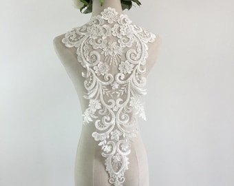 Puede incluir: Una aplicación de encaje blanco con un intrincado diseño floral y de volutas. La aplicación se muestra en un maniquí, mostrando sus delicados detalles y su potencial para adornar ropa de novia o formal. El diseño presenta patrones simétricos y una variedad de motivos florales.