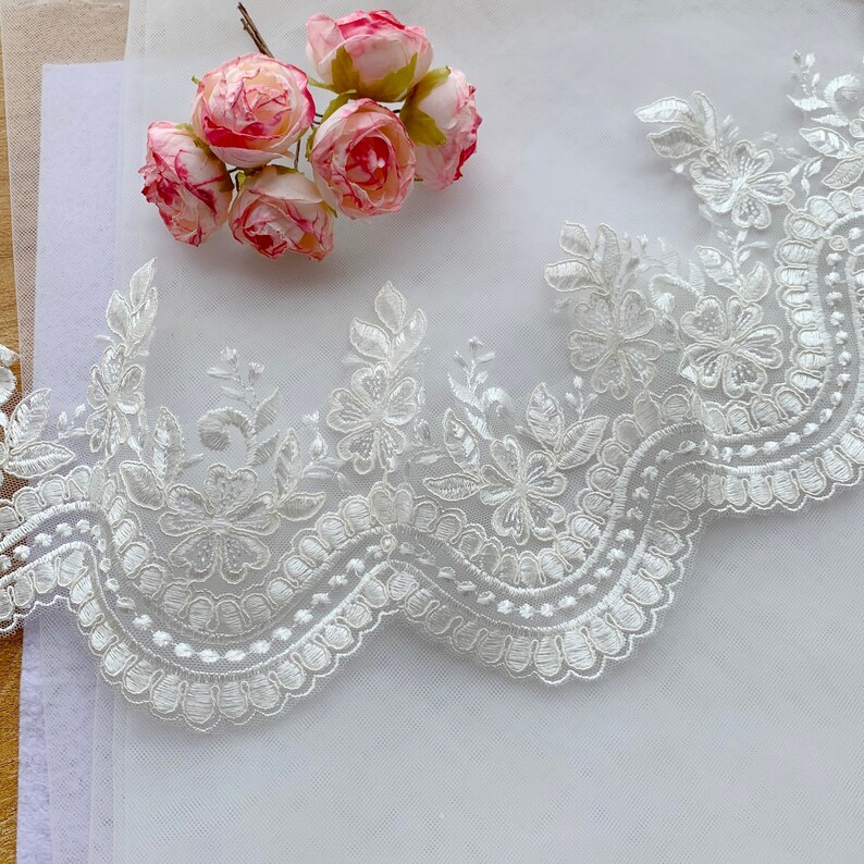 Puede incluir: Ribete de encaje floral blanco con borde festoneado, con flores y hojas bordadas. Un peque&ntilde;o ramo de rosas rosas y blancas descansa cerca. Ideal para proyectos nupciales o de artesan&iacute;a.